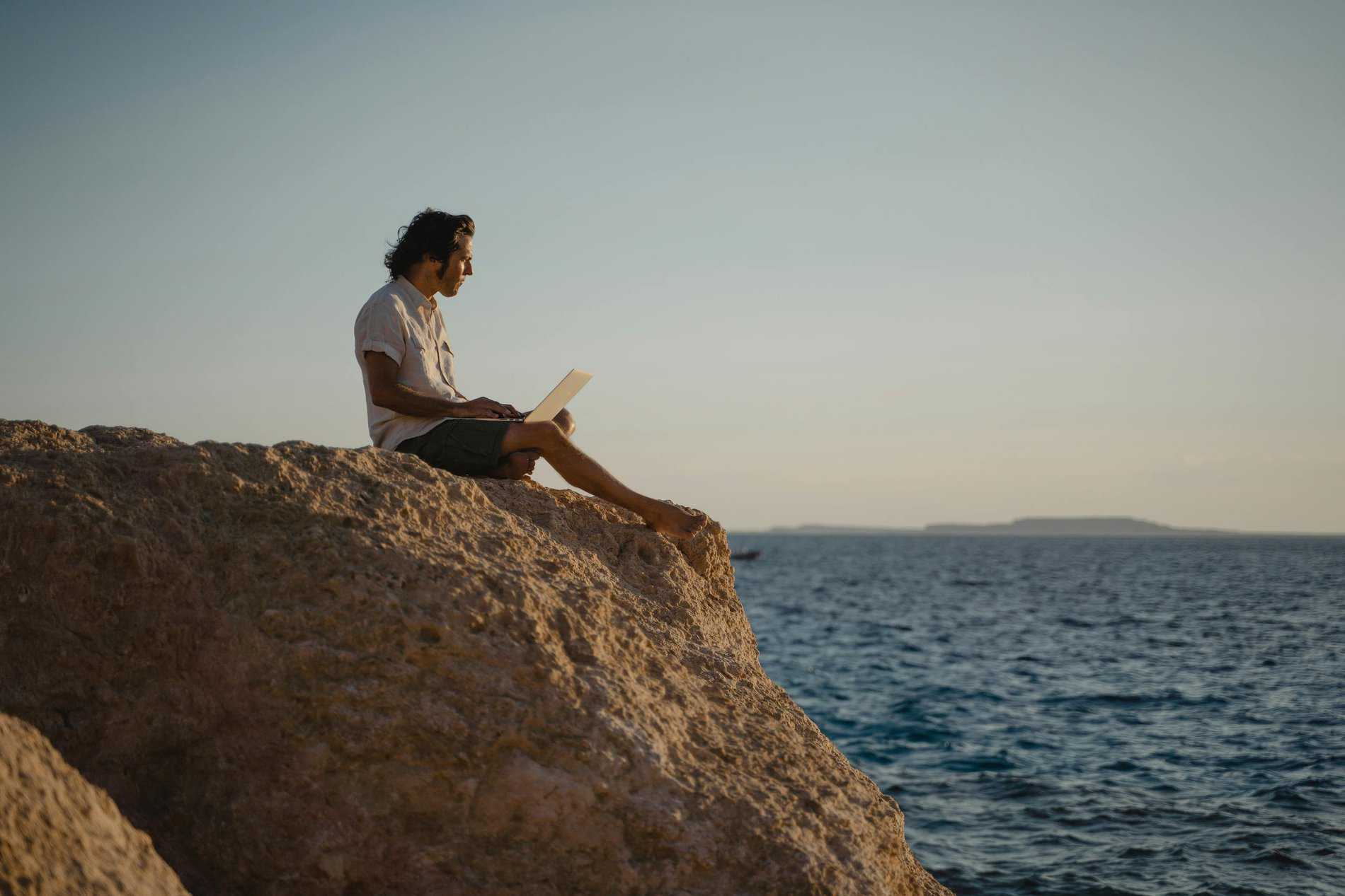 Man in White Shirt Sitting on Brown Rock Formation Near Body of Water